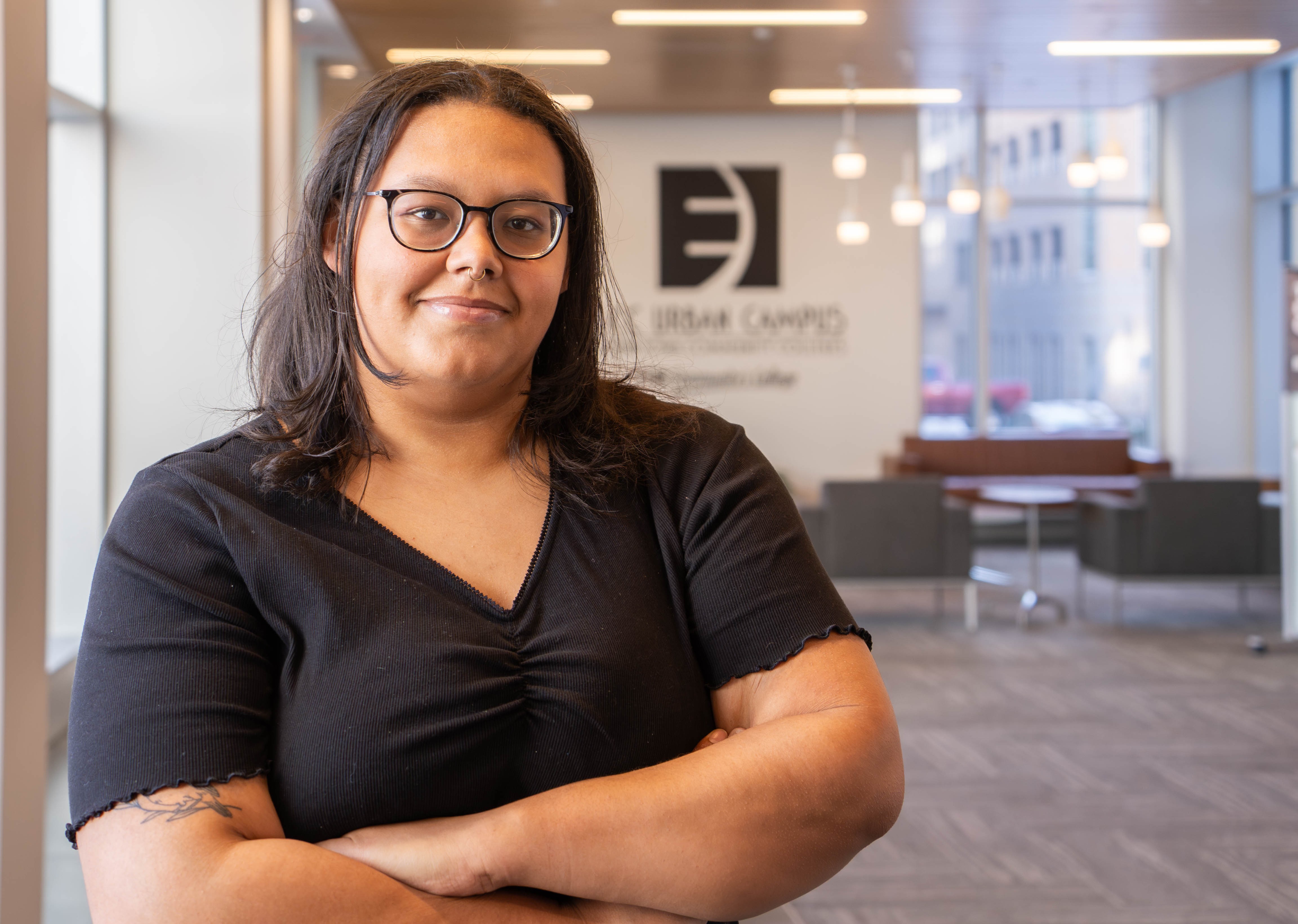 Harli Harrington, EICC alumna, stands indoors with arms crossed, smiling slightly. She wears glasses and a black top, with a softly lit campus lobby and EICC signage in the background.