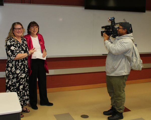 EICC's Muscatine campus President Naomi DeWinter stands with Associate Professor Lori Sheppeard, both facing a WQAD camera operator who is on-site to record the presentation of the Pay It Forward award.  
