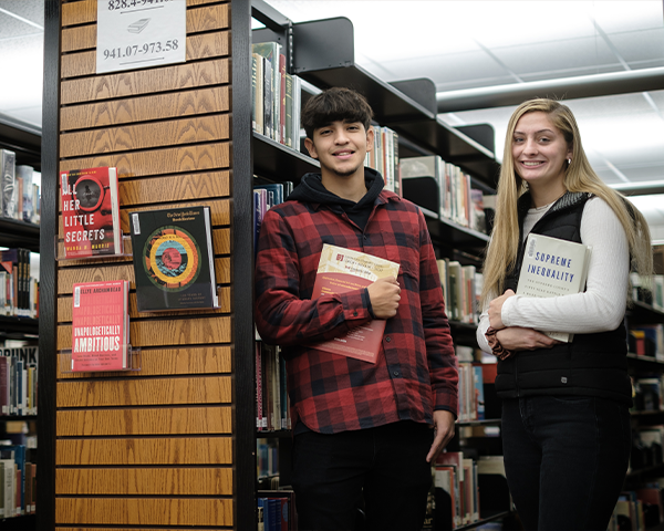 Two students standing amoung bookshelves smiling holding books