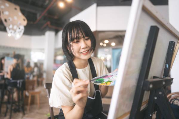 smiling woman in painter overalls sitting at an easel painting 